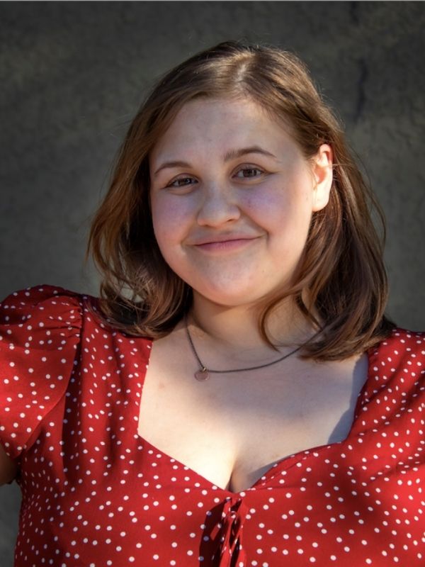 Young woman smiling. Medium length brown hair is loose. She wears a gold nacklace and a red dress with white dots