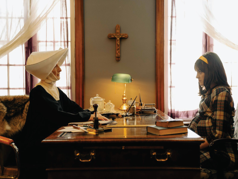 Film still with two women sitting across from each other on a desk. The room is brown and seldom