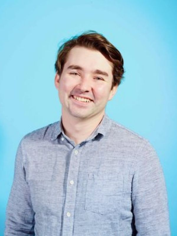 Young man with brown short wavy hair smiling and wearing a blue button-up collared shirt