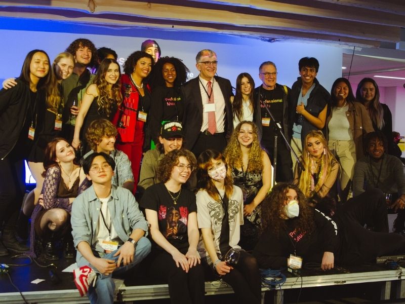 A group of students and faculty standing on a brightly lit stage smile for a camera