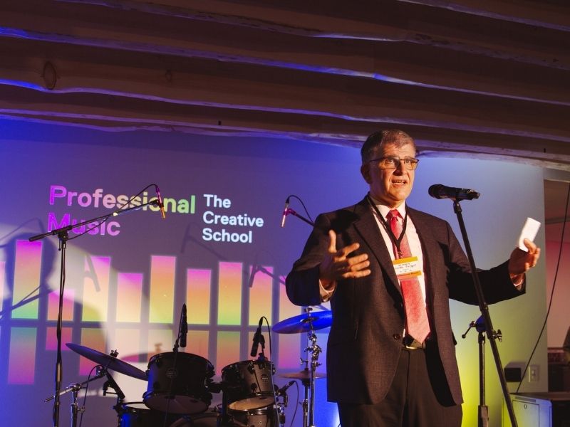 A man wearing a dark suit with a red tie standing on a stage and speaking into a microphone. The stage is brightly lit with a drum set in the background. 