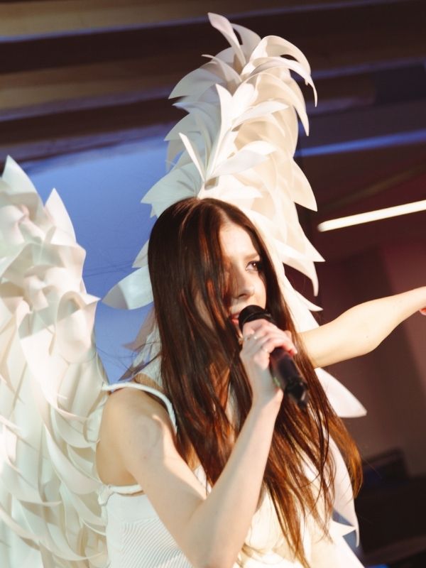 A girl singing into a microphone wearing all white with two large, white angel wings clipped onto her back.