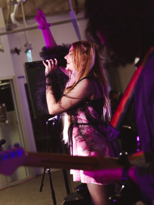  A man with long hair plays bass and faces away from the camera in the foreground. A girl in the middle ground is singing into a microphone and holding her left arm up.