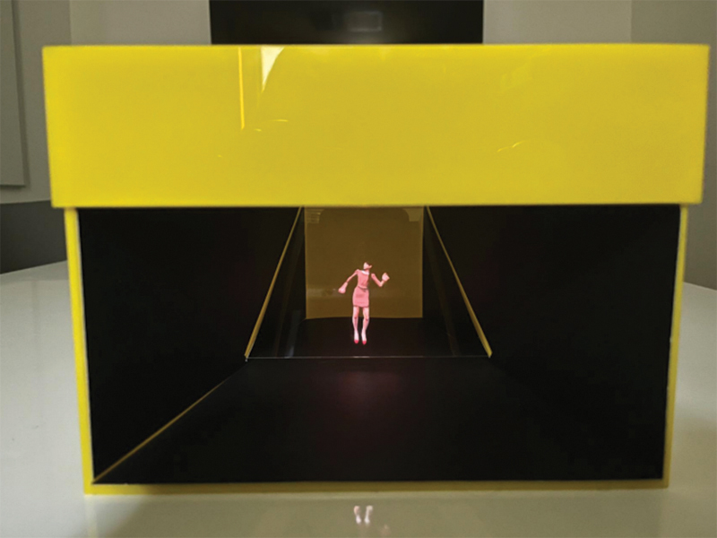 A constructed box with a panel of glass resting inside at a 45-degree angle. A beam of light refracts off of the glass, creating a holographic render of a human