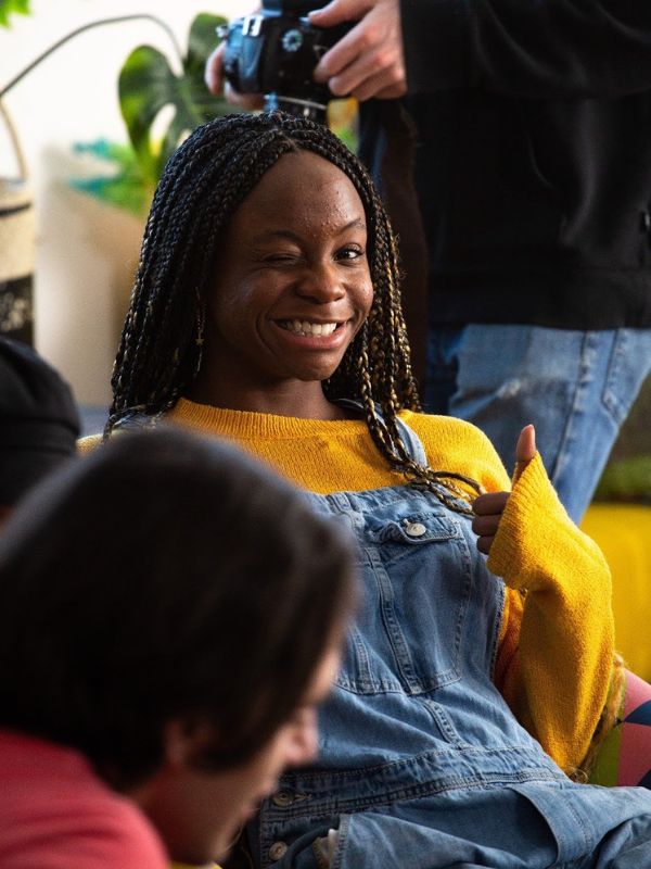 A girl wearing a yellow sweater and blue overalls smiling towards the camera and holding a thumbs up. 