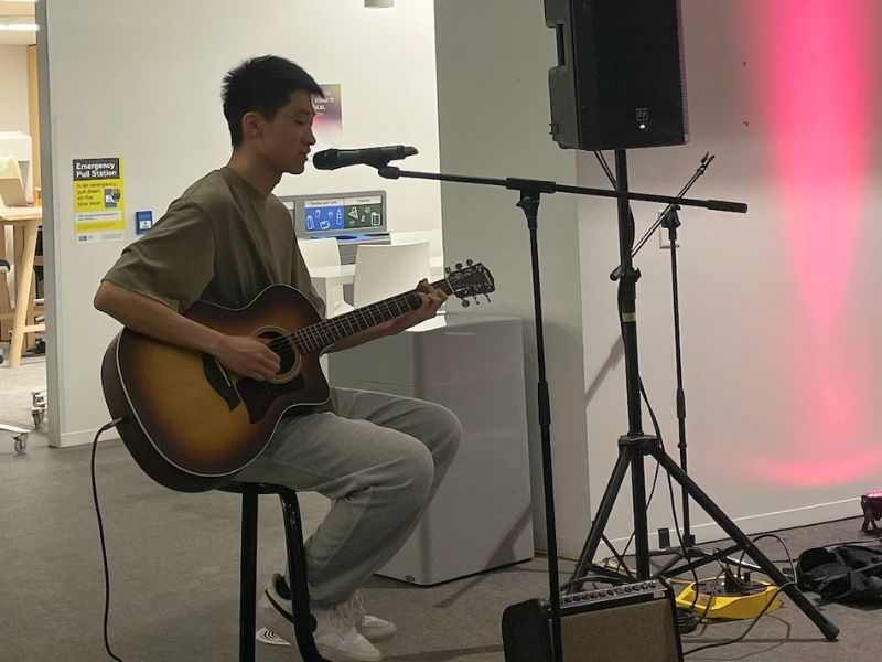 Liam Hao has short black hair. He sits on a stool, playing an acoustic guitar while singing into a microphone.