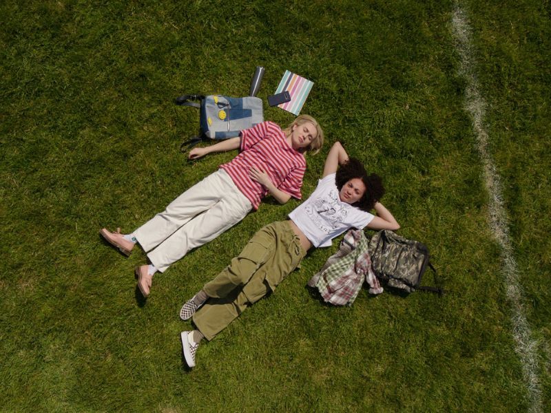 Two women laying down in a grassy green field looking up at the cloud. Books and backpacks are also resting in the grass beside them.