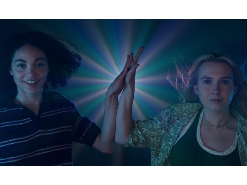  Two women standing tall look assuredly into the distance. Their hands are high fiving in the middle of the frame with an iridescent glow behind them illuminating the otherwise dark background.