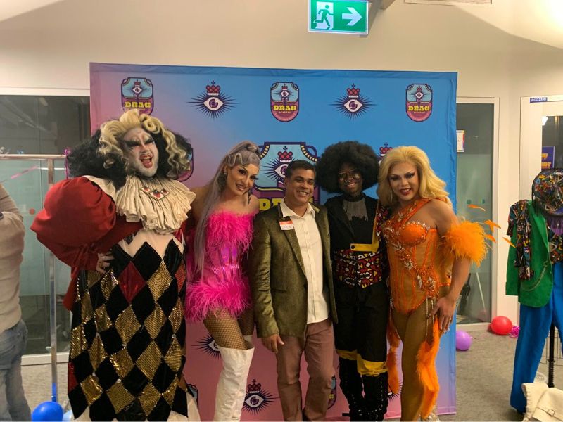 Five people dressed in drag attire posing in front of a Drag Academy backdrop.