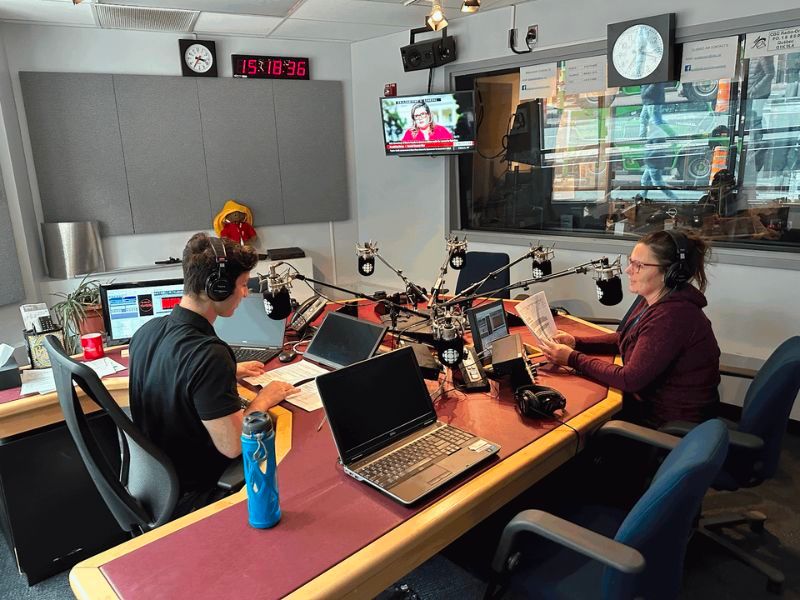 A man and a women wearing headphones and sitting in a broadcast room, appearing to be working and speaking into microphones