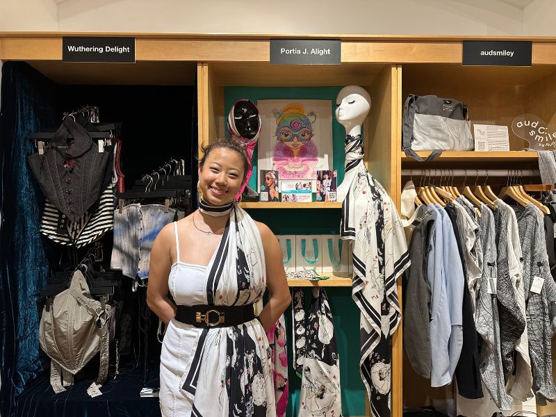 Portia J. Alight is posing in front of her Italian silk scarf collections. She is wearing a white dress, black and gold belt, and one of her scarves around her neck and tucked through the belt.