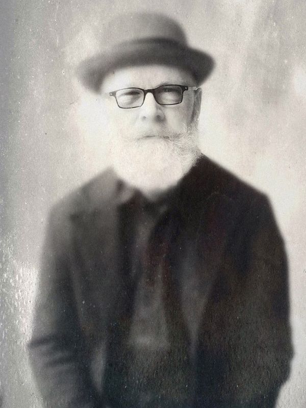 A black and white image of a man wearing a black suit, glasses and brimmed hat stares stoically at the camera