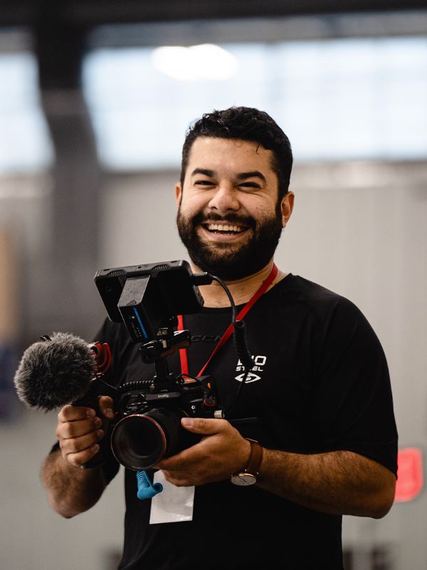 A man posing holds a professional camera and smiles at the camera 