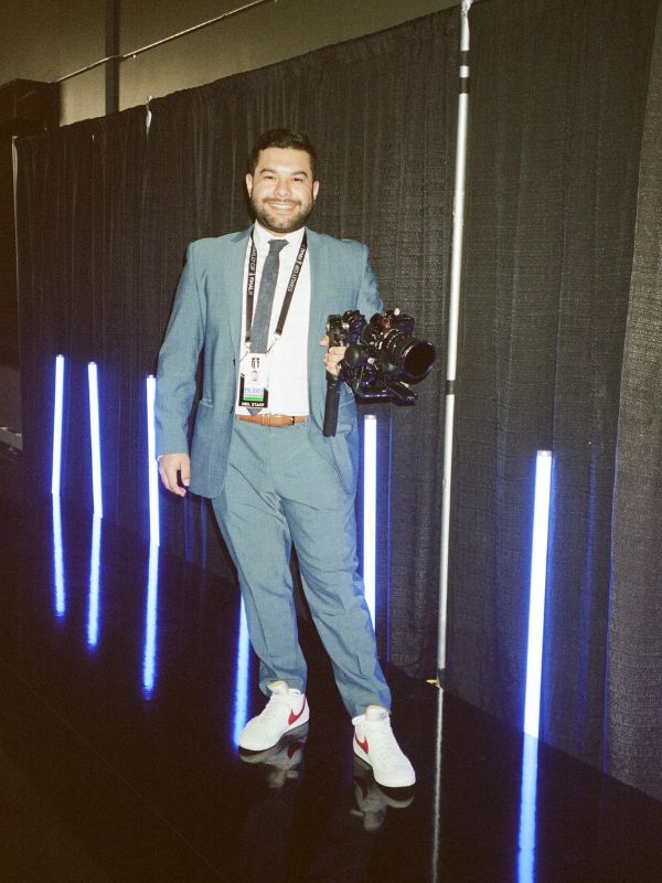 A man posing in a light grey suit holds a professional camera on a tripod and smiles at the camera 